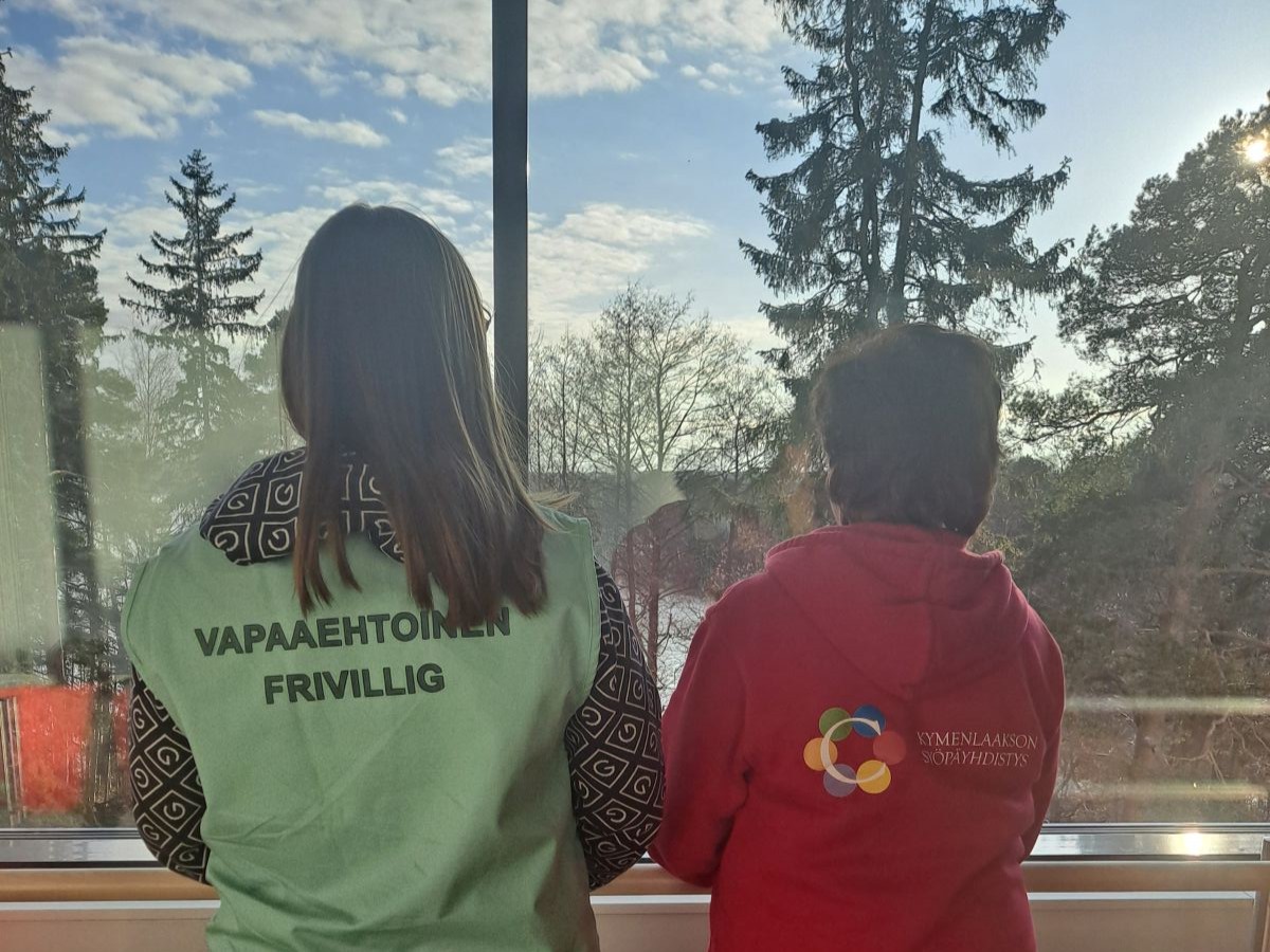 Kaksi ihmistä seisoo ikkunan edessä, selät kameraan päin. Vasemman puoleisella henkilöllä on yllään OLKAn liivi, jonka selässä lukee "vapaaehtoinen - frivillig". Oikean puoleisella henkilöllä on yllään punainen huppari, jonka selässä lukee "Kymenlaakson Syöpäyhdistys".