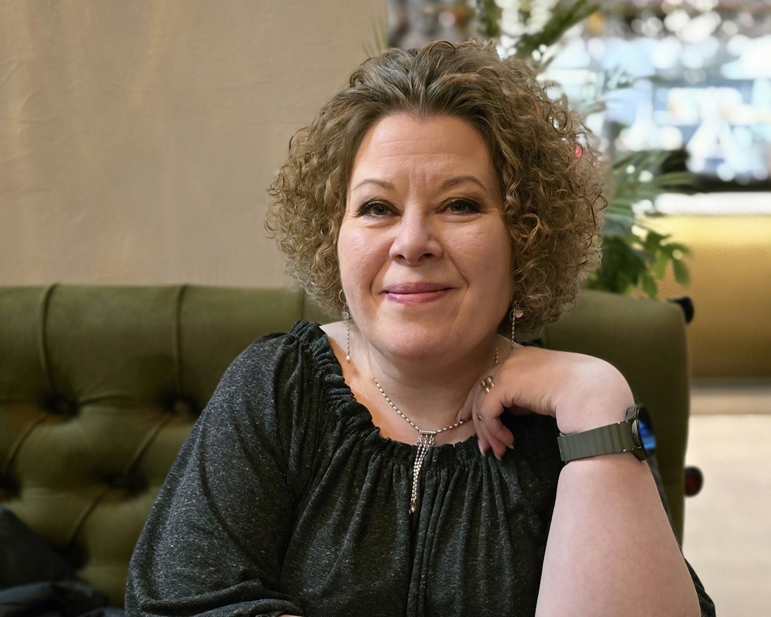 Kuva Soilesta istumassa kahvilapöydän ääressä. Soile katsoo hymyillen kameraan, hänellä on päällään tumma, kimalteleva paita. Taustalla näkyy myös vihreää sohvaa.