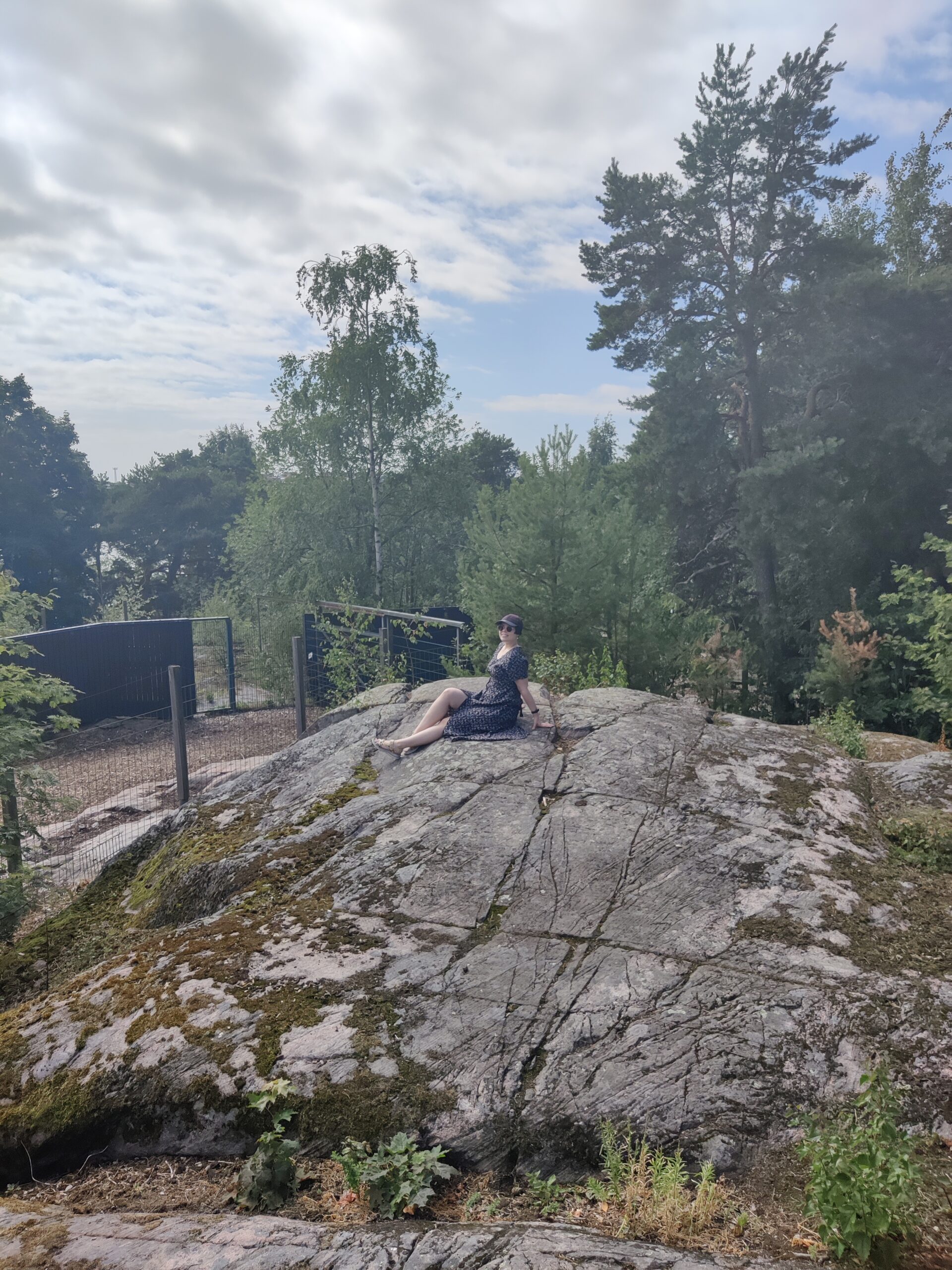 Anita istumassa kalliolla, katsoen hymyillen kameraan. Anitan taustalla näkyy metsäistä lehtipuumaisemaa.
