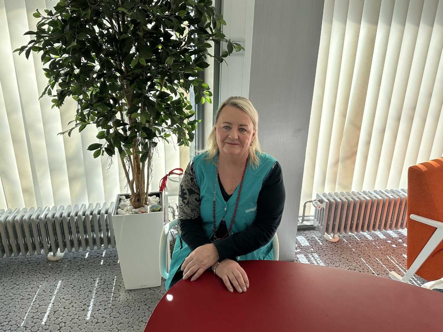 Vapaaehtoisena toimiva Eila istuu punaisen pöydän ääressä. Eila hymyilee kameralle ja hänellä on yllään vaaleanvihreä vapaaehtoisen liivi.