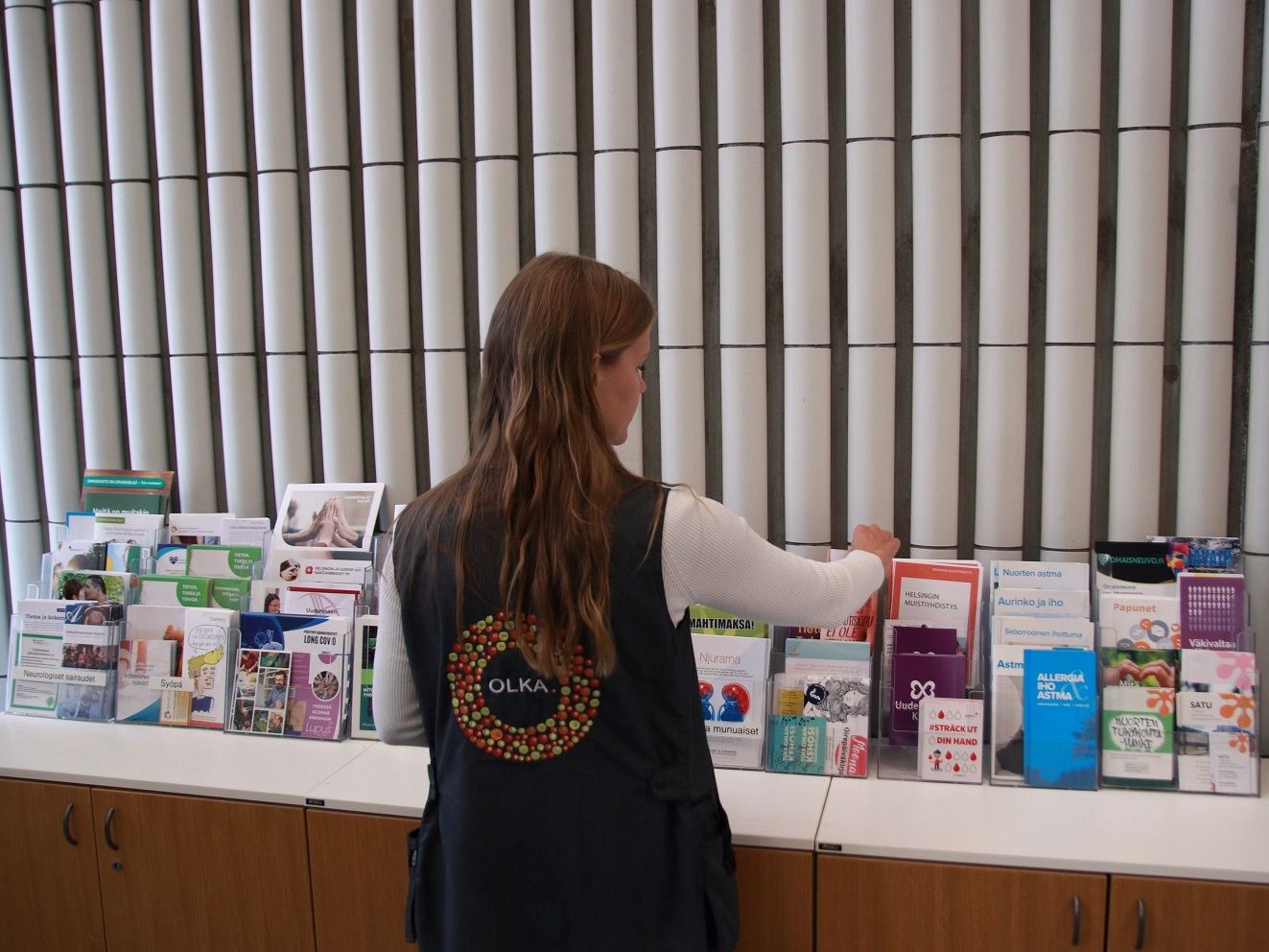 OLKAn työntekijä järjestelemässä esitteitä OLKA-pisteellä. Hän on selin kameraan, ja päällään hänellä on harmaa liivi, jonka selässä on OLKAn logo.