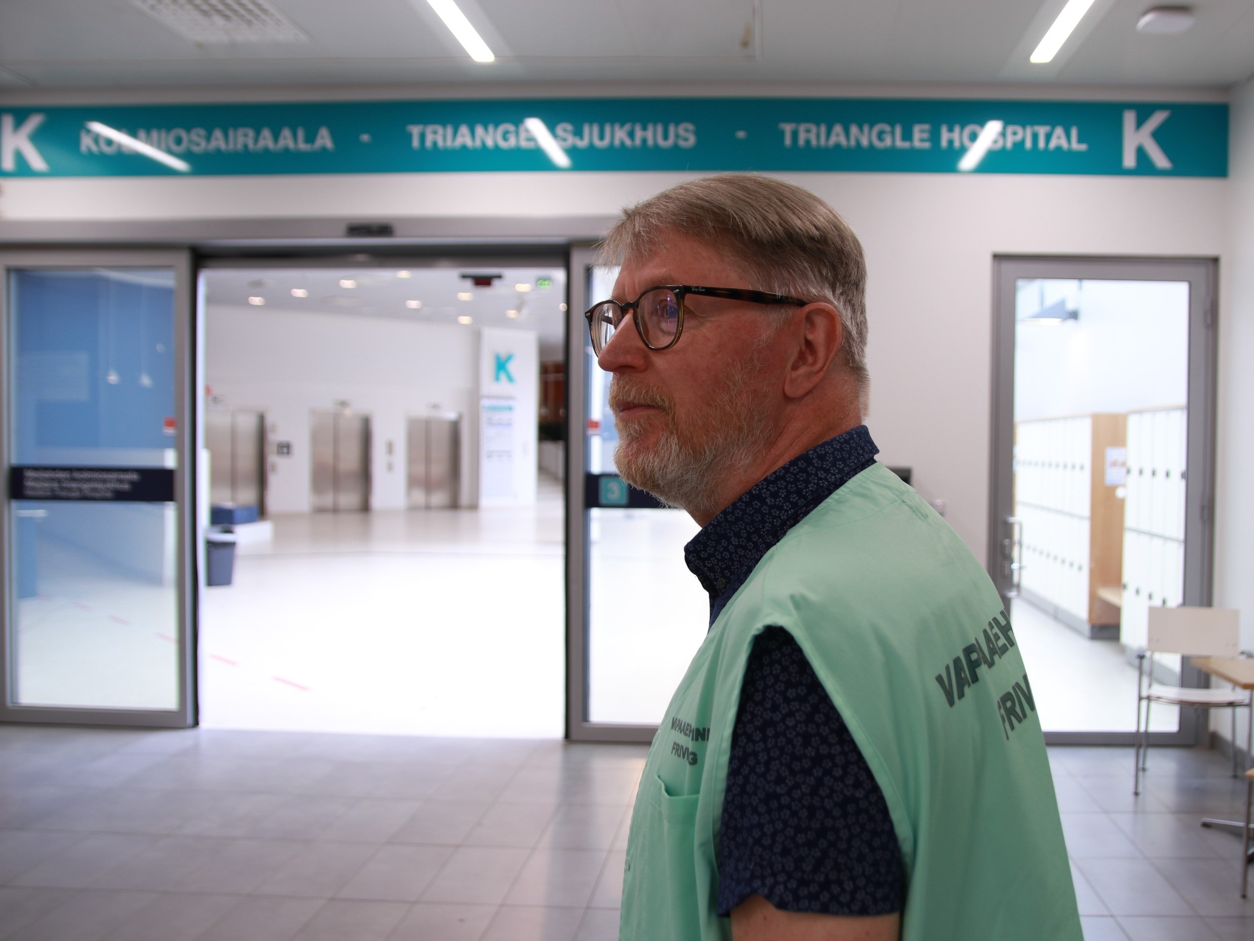 OLKAn vapaaehtoinen Meilahden sairaaloiden pääaulassa. Vapaaehtoinen katsoo pääoville päin, valmiina auttamaan. Hänellä on yllään vaaleanvihreä vapaaehtoisen liivi.