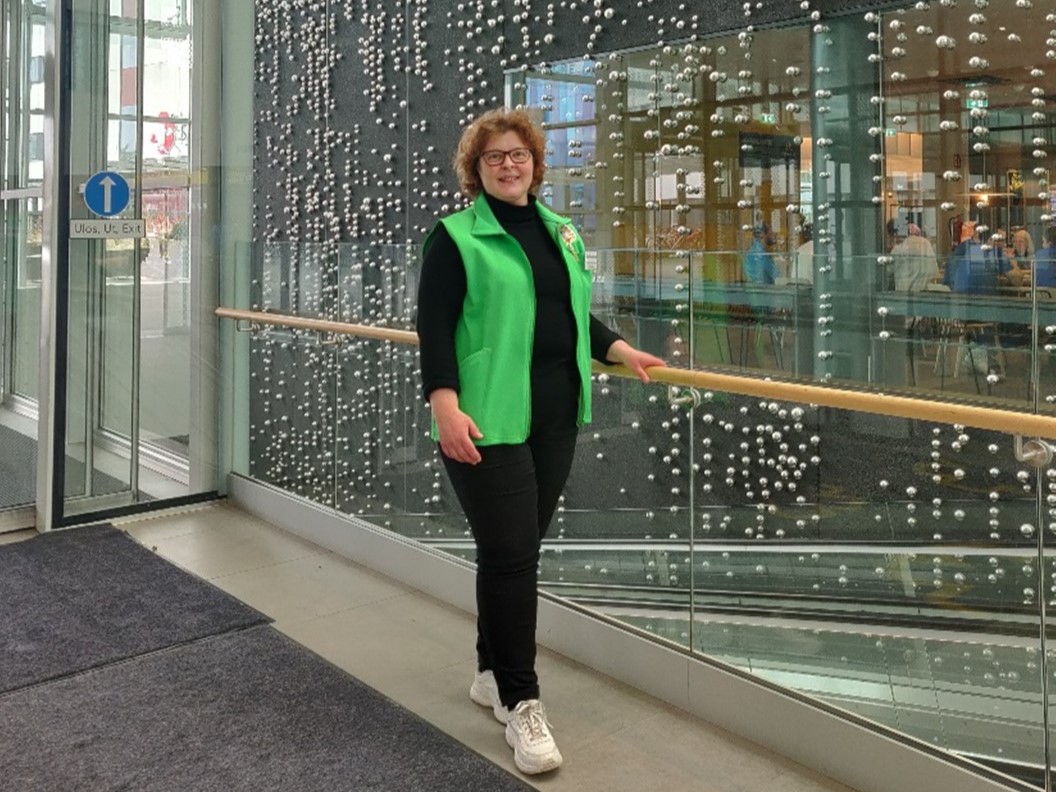 Pirkanmaan OLKAn vapaaehtoinen Johanna hymyilee vapaaehtoisen vihreä liivi päällään. Johanna seisoo korkean seinän edessä, johon on kiinnitetty koristeeksi hopean värisiä metallipalloja pitkiksi jonoiksi.
