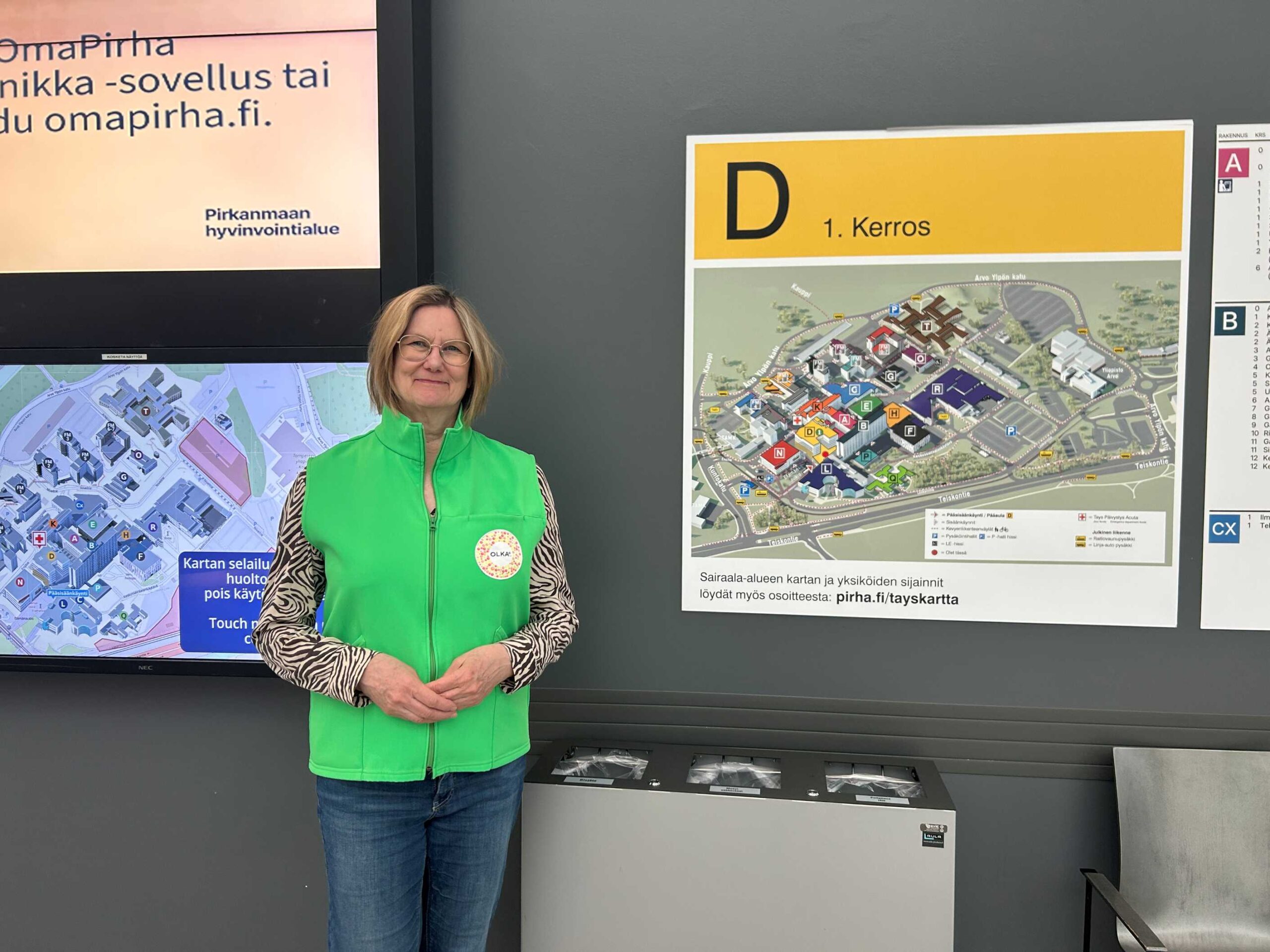 OLKA Pirkanmaan vapaaehtoinen Kristiina sairaalakartan edessä. Kristiina hymyilee kameralle, ja päällään hänellä on vapaaehtoisen vihreä liivi.