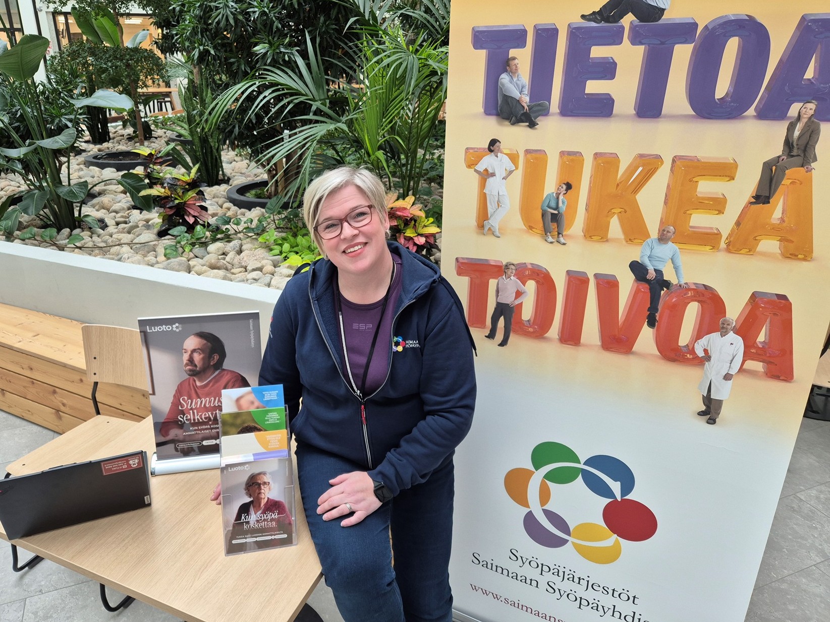 Terveydenhoitaja Janna Puurunen OLKA-pisteellä, esittelemässä Saimaan Syöpäyhdistyksen toimintaa. Janna katsoo hymyillen kameraan ja hänen ympärillään on erilaisia esitteitä ja yhdistyksen roll up -mainos.