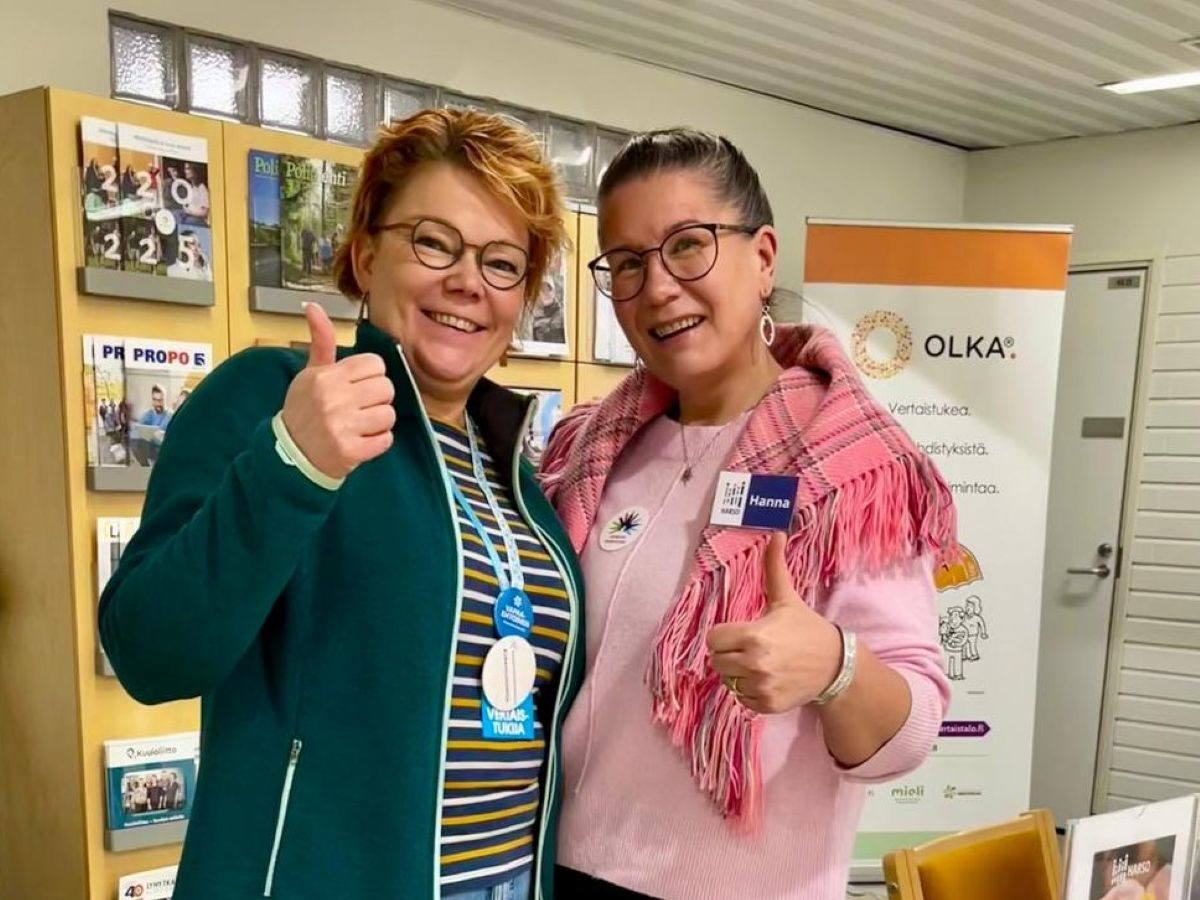 Kaksi naishenkilöä seisoo OLKA-pisteen edessä, hymyilee kohti kameraa ja näyttää peukkuja. He näyttävät iloisilta.