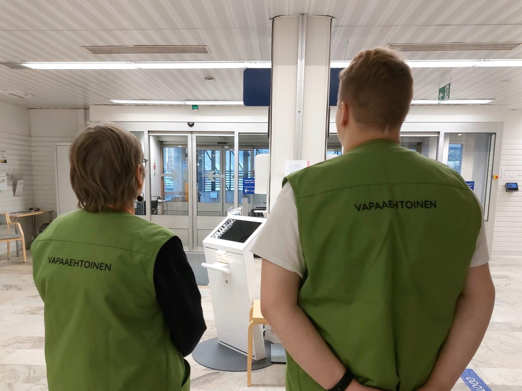 Kaksi OLKA Lapin vapaaehtoista vapaaehtoisten vihreät liivit yllään. Vapaaehtoiset seisovat selin kameraan, ja taustalla näkyy sairaalan aula ilmoittautumisautomaatteineen.