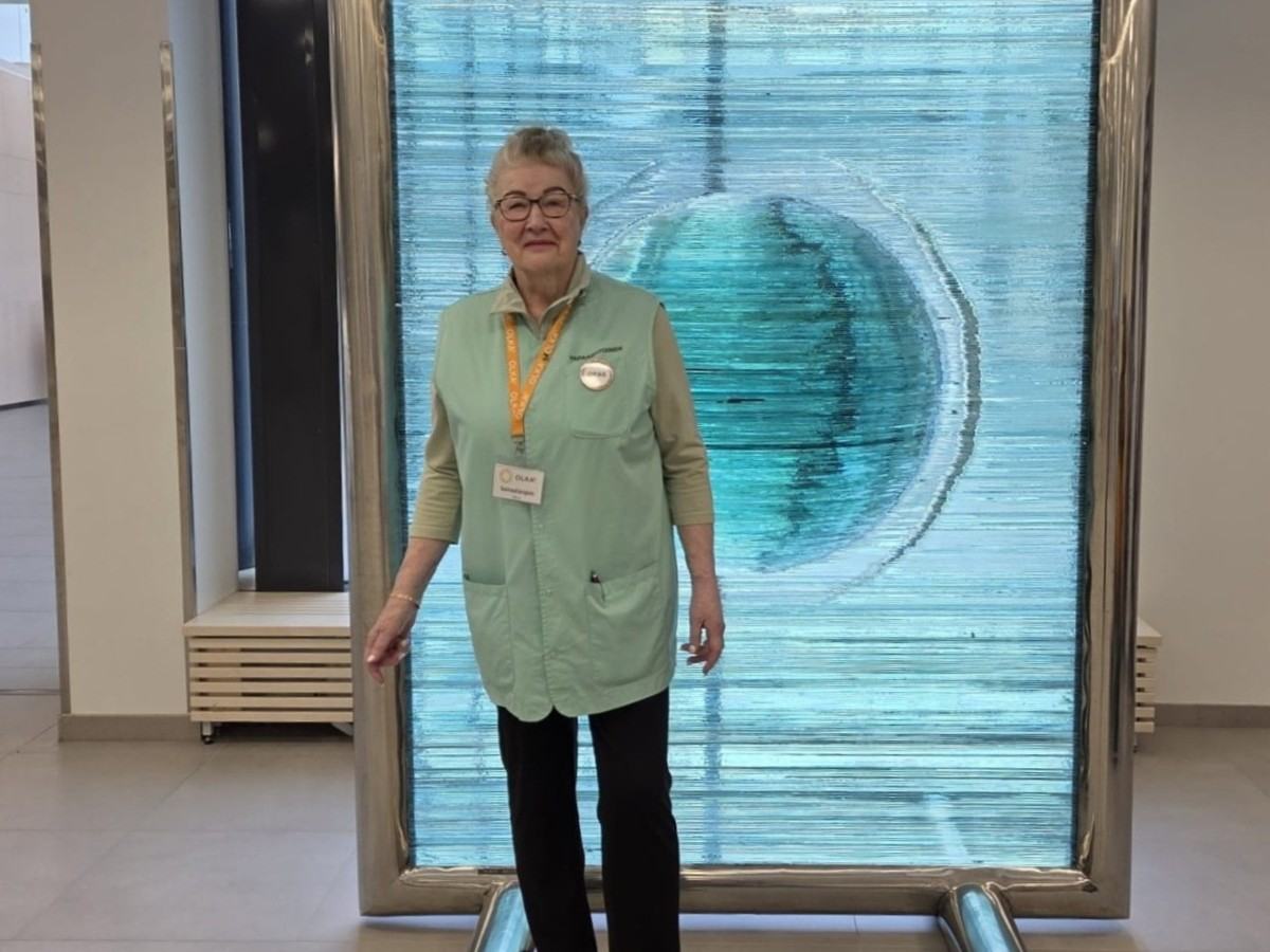 Päijät-Hämeen OLKAn vapaaehtoinen Ritva sairaalan aulassa. Ritvalla on yllään vapaaehtoisen vaaleanvihreä liivi, ja hän katsoo kohti kameraa hymyillen.