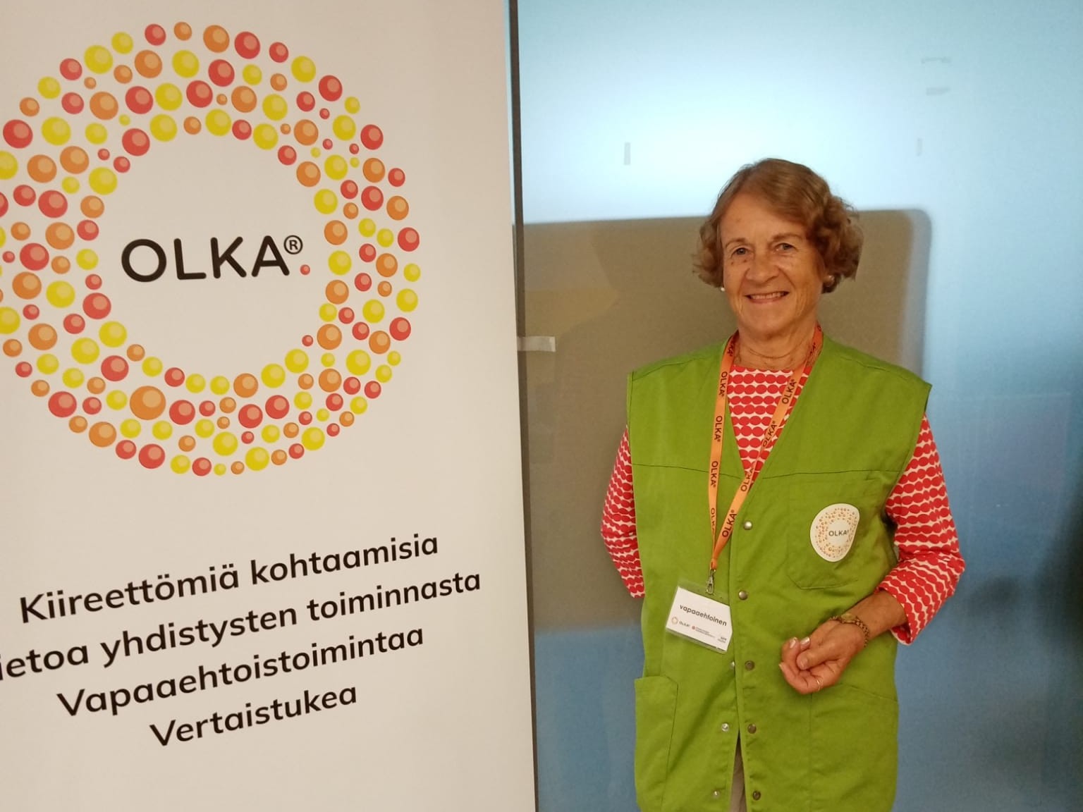 OLKA Pohjois-Karjalan vapaaehtoinen OLKA-pisteellä. Ulla hymyilee kameralle, ja hänellä on päällään vapaaehtoisen vaaleanvihreä liivi.