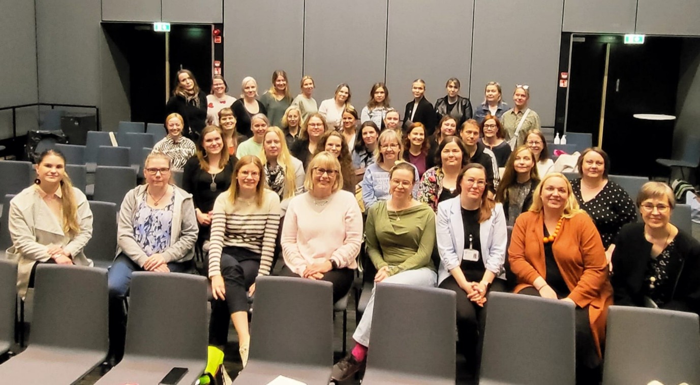 Valtakunnallisen OLKA-verkoston työntekijät OLKAimissa 2025. Työntekijät istuvat Sairaala Novan luentosalissa ja katsovat hymyillen kameraan.