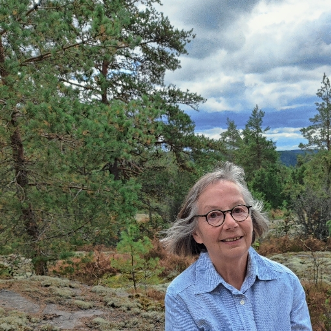 Vertaistukija Riitta istumassa hymyillen kalliolla, metsäinen maisema takanaan. Riitalla on sininen paitapusero ja tummasankaiset silmälasit.