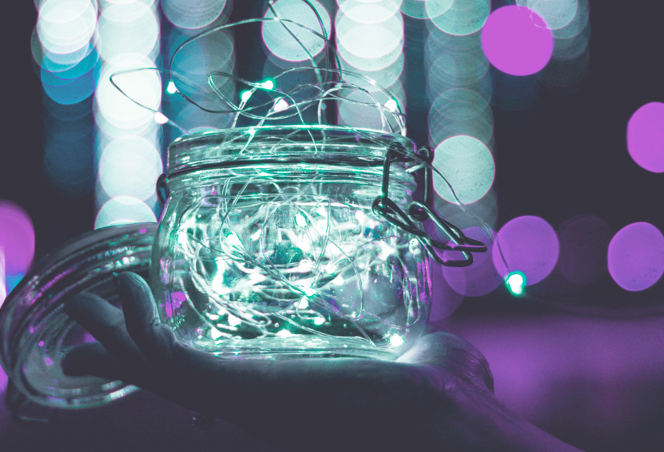 Käsi, joka kannattelee lasista purkkia. Purkin sisällä ja ulkopuolella on pieniä, vaaleita valoja. Taustalla näkuu turkoosin ja violetin eri sävyjä.