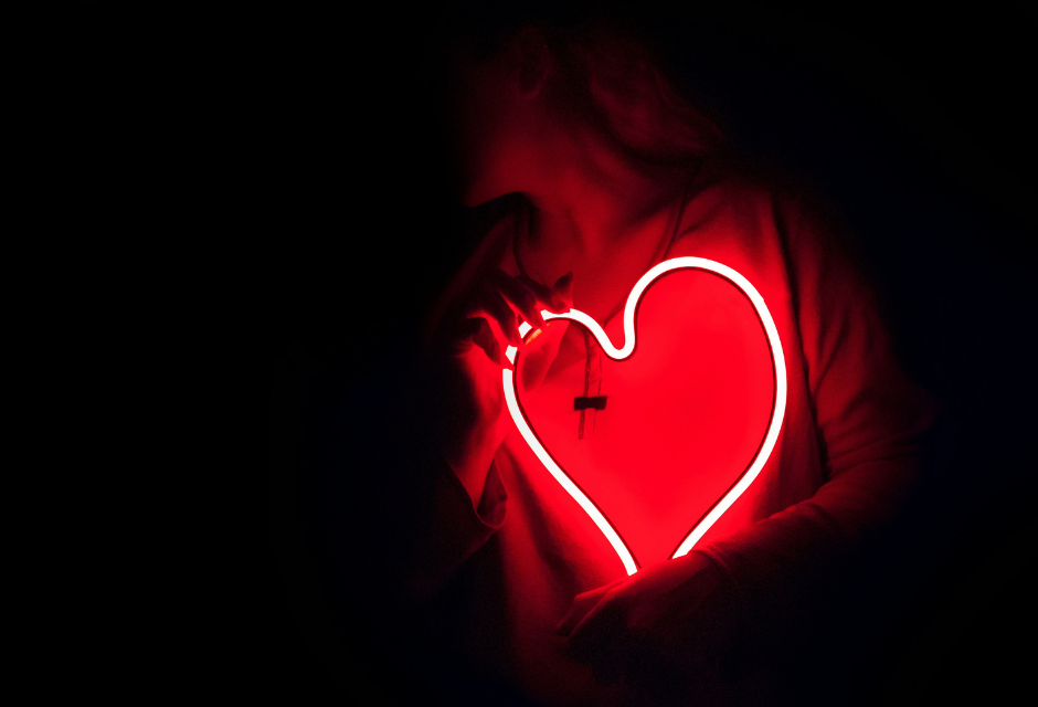 Ihminen täysin pimeässä tilassa, kasvot osittain varjossa. Henkilöllä on sylissään punainen led-valo sydän, joka heijastaa hieman valoa hänen ylleen.