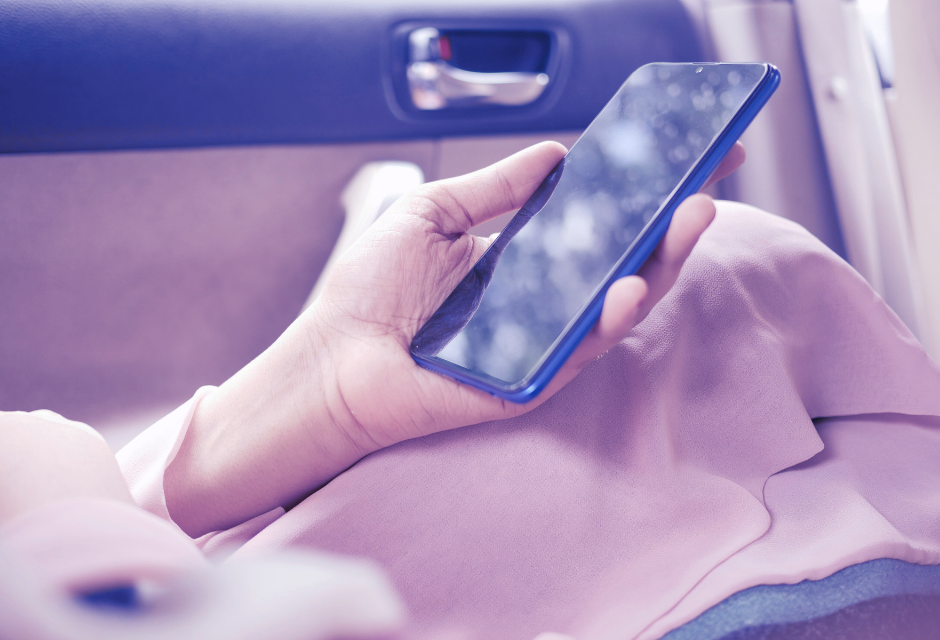 Naisen käsi jalkaa vasten, autossa. Taustalla näkyy auton sisäpuolelta oven kahva. Kädessä on älypuhelin.