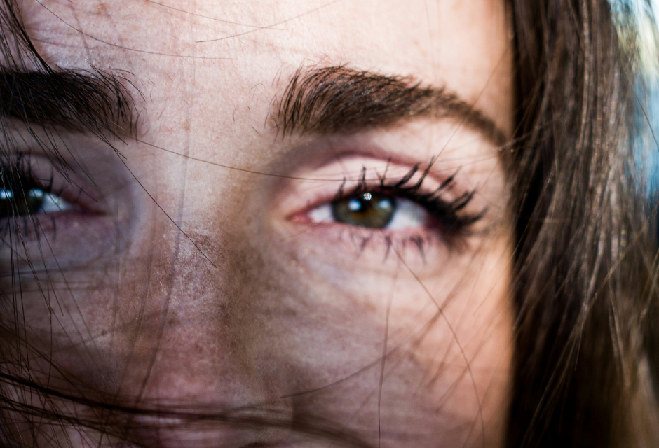 Lähikuva naisen kasvoista. Nainen katsoo kohti kameraa ja hänen kasvoillaan on hiuksia. Taustalla kaukaisuudessa näkyy ulkomaisema.