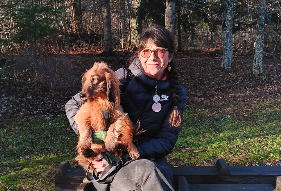 Saara Suvitie istumassa metsän laidalla penkillä. Aurinko paistaa ja maisema on syksyinen, Saaralla on sylissään pieni koira.