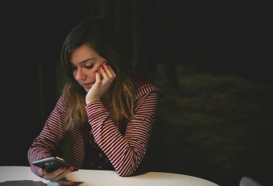 Tummahiuksinen nainen istuu pöydän ääressä ja katselee kädessään olevaa älypuhelinta, nojaten leukaansa toiseen kämmeneensä.
