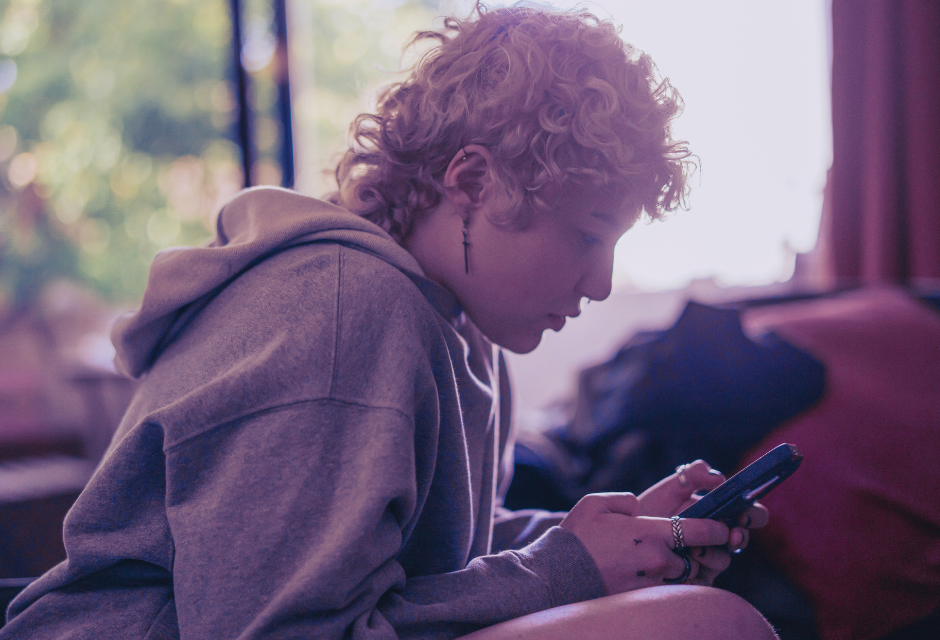 Muunsukupuolinen henkilö katselemassa puhelintaan huoneessa, jonka ikkunasta paistaa aurinko.