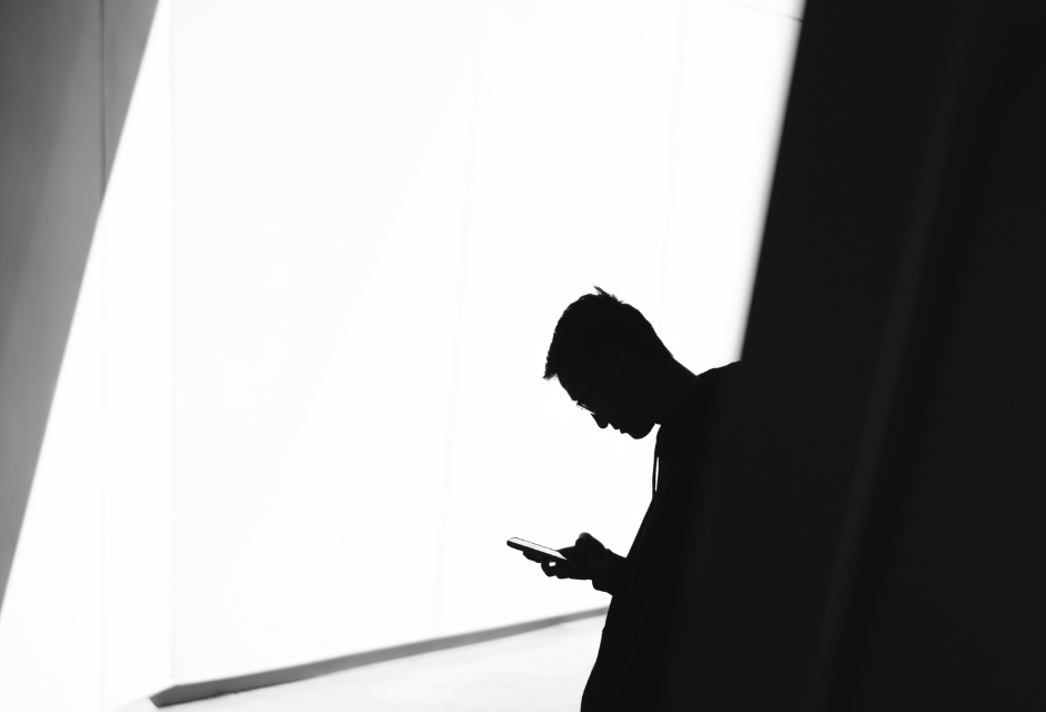 Mieshenkilö pitää puhelinta kädessään ja katsoo sitä, pää alaviistoon taitettuna. Mies jää mustavalkoisessa kuvassa varjoon siten, että häntä ei voi tunnistaa.
