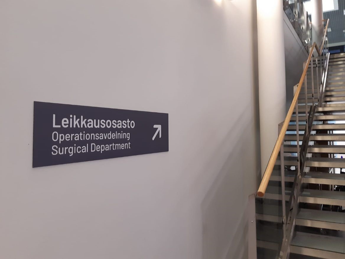 Kuva sairaalan opastekyltistä seinällä. Opastekyltissä lukee "Leikkausosasto" ja nuoli osoittaa rappusia ylöspäin.