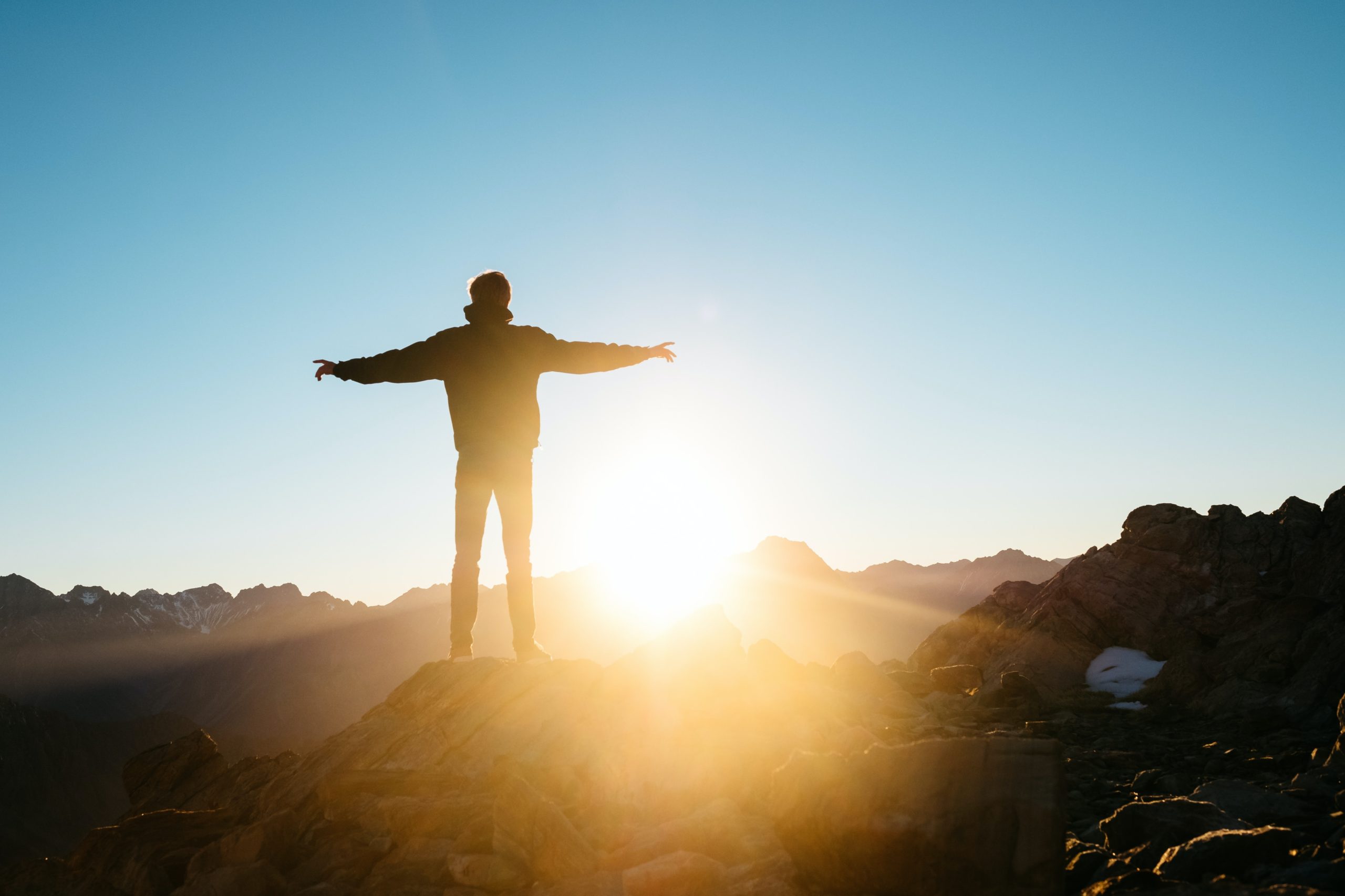 Henkilö seisoo vuoristossa kohti aurinkoa, kädet sivuille ojennettuina